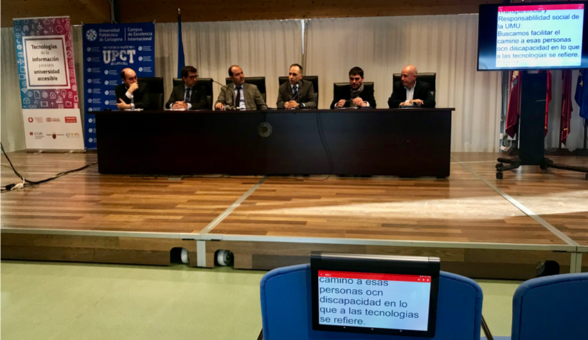 Se ve una mesa de ponentes en un congreso, un monitor con la transcripción simultánea de Ability Connect y una tablet en la que también se está recibiendo el texto