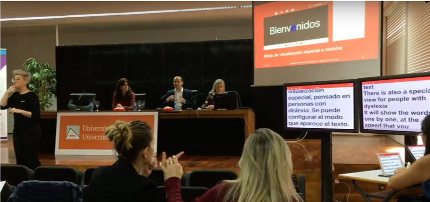 Una mesa de ponentes en una jornada donde se ven dos monitores con la trasncripción de lo que ocurre en castellano e inglés con Ability Connect